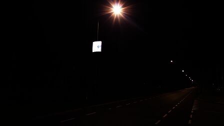 Terschuur, weg in de avond met lantaarnpalen. Aan 1 lantaarnpaal hangt een bord met verlichte reclame. Daar richt je oog zich op