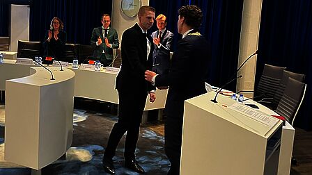 In de raadszaal schudt de burgemeester de hand van John van Ginkel om hem te feliciteren. De andere aanwezigen klappen.
