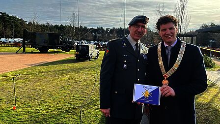 De commandant en de burgemeester staan buiten ter gelegenheid van het 150-jarig bestaan van de Verbindingstroepen. Op de achtergrond staan militaire voertuigen.